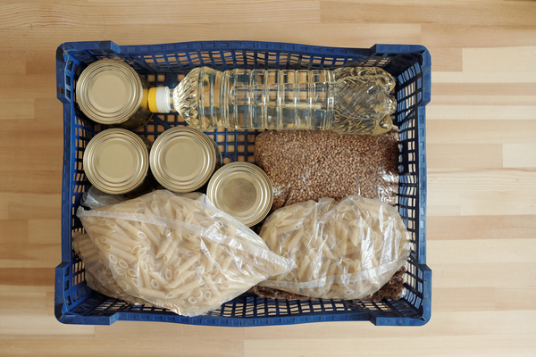 Silver cans cellophane bags with penne bags with buckwheat groats and a bottle of oil are in an indigo plastic box that is located on a light wood surface