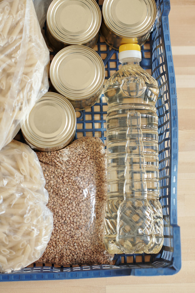 A bottle of oil is placed in plastic crate with canned goods pasta in cellophane packets and a buckwheat package