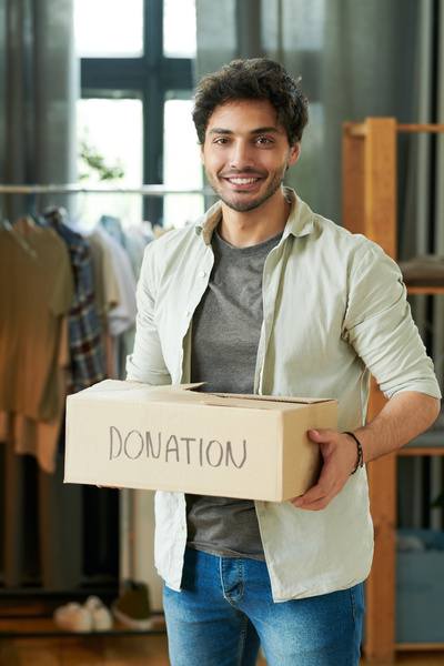 Smiling black haired man with bristles wearing ivory shirt and jeans holds charity box in a room