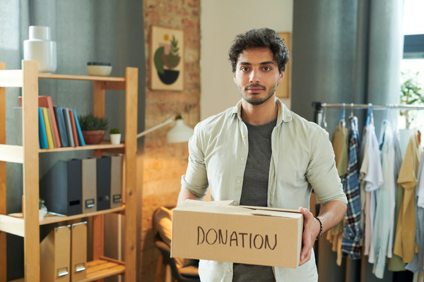 A man with dark hair in ivory shirt holds a box with an inscription about charity and with donations inside