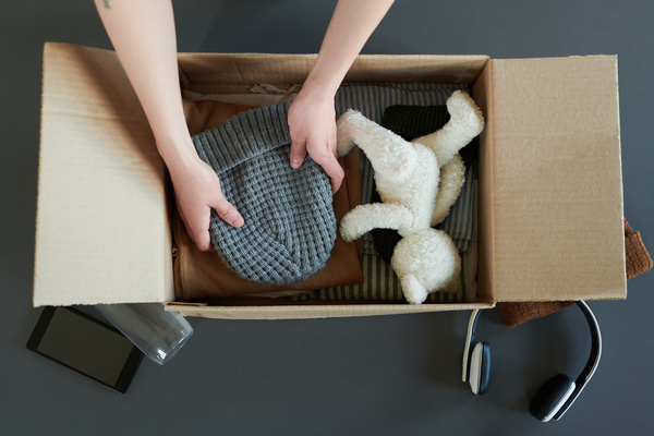 A man packs a stack clothes and gray knitted hat in a box with white stuffed animal and striped items of clothes which is on the surface of dark grey with smartphone a bottle brown hat and black-white headphones near