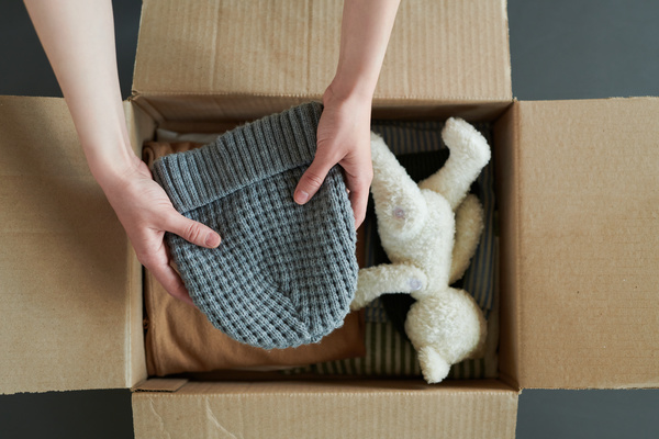 A man packs a stack clothes and gray knitted hat in a box with white stuffed animal and striped items of clothes which is on the surface of dark grey