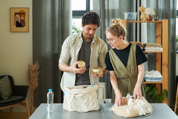A blonde wife with round glasses and a husband with bristles put products in light colores string bag and canvas tote which are on the table with a bottle of water near in a bright room with a wooden rack