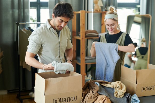 A black haired man with bristles and a woman with round glasses and a headband fold and put clothes in boxes boxes with lots of clothes between them