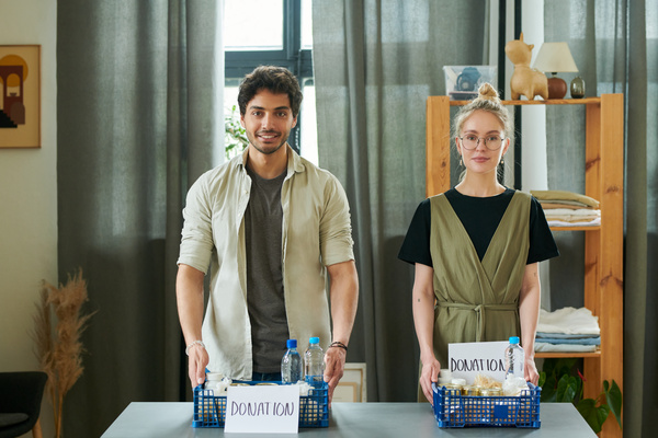 A man and a woman in casual clothes have their hands on indigo colored plastic donation crates full of products with sheets of paper on which it is written about the donation