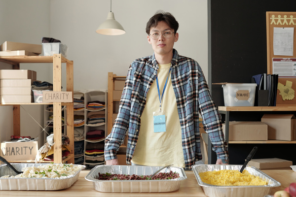 Male volunteer with glasses in checkered shirt and light colored jeans stands behind the table with food in aluminum forms in a room with wooden racks and boxes