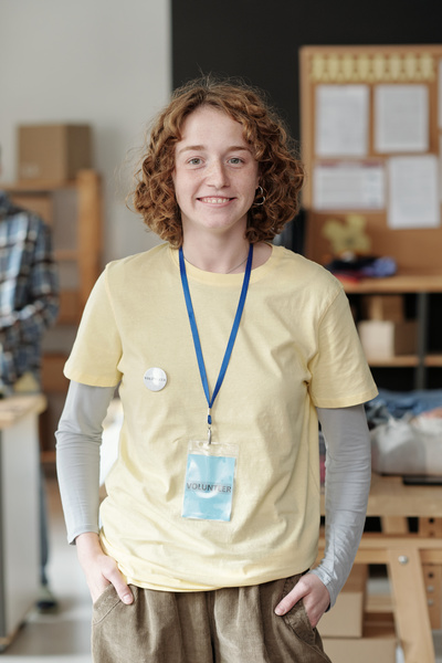 Smiling female volunteer with red curly hair dressed in light colored shirt over white longsleeve stands with her hands in pants pockets