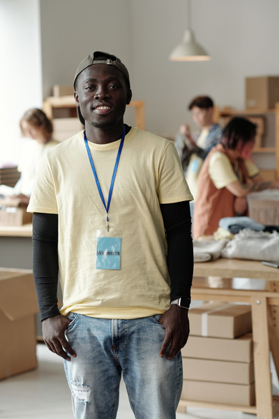 Young smiling voluntary worker with a cap and a blue badge dressed in light yellow t-shirt over black longsleeve and a jeans stands with his hands in his jeans pockets