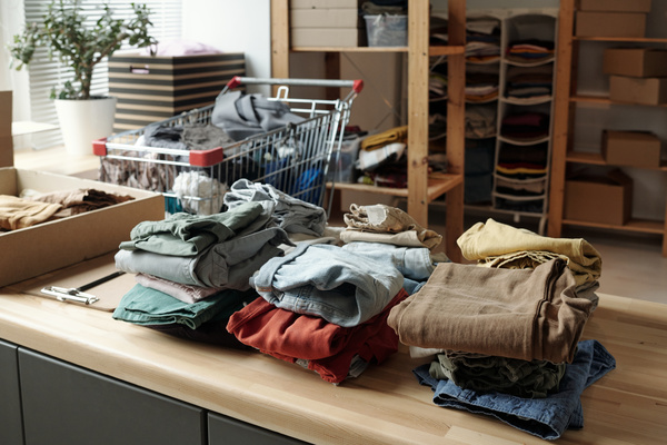 Stacks of folded clothes of different colors are placed on the table next to a box with clothes in it in a room with wooden racks