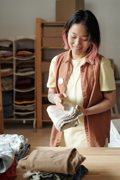 Smiling asian voluntary worker with dyed bob haircut dressed in a coral sleeveless blouse over ivory t-shirt folds denim piece of clothing standing behind the table with stacked clothes