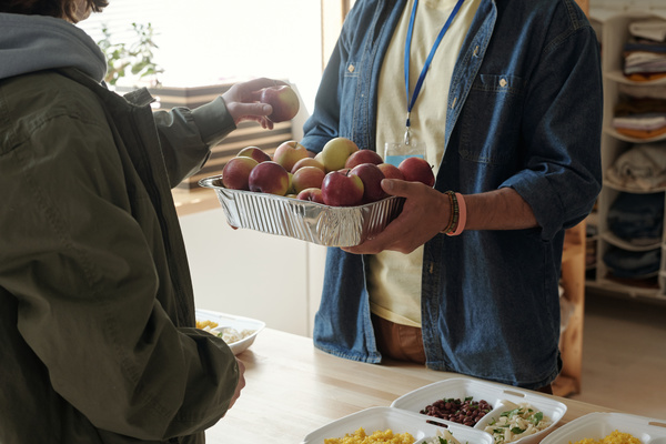 Voluntary worker with bracelets and a blue badge in denim shirt over pale yellow t-shirt gives apples from an aluminum form to the person in need