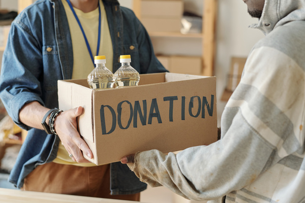 Volunteer with bracelets and digital watch in denim shirt over pale yellow t-shirt gives a box with two bottles of oil on which there is an inscription about donation