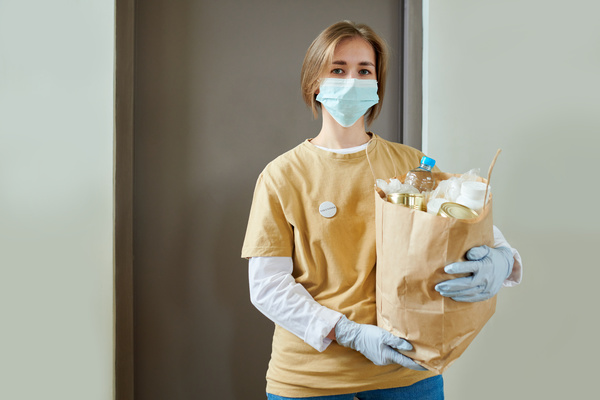 Female volunteer in medical mask with short hair and button badge on her yellow t-shirt with two hands in rubber gloves holds a craft package with products standing at the grey door