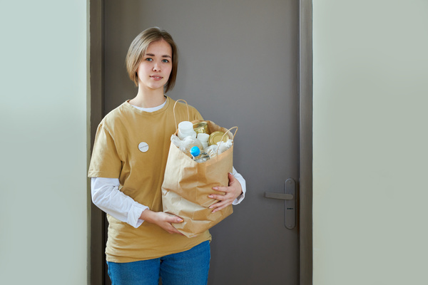 Female volunteer with short hair and button badge on her yellow t-shirt stands with a craft package full of products at the gray door