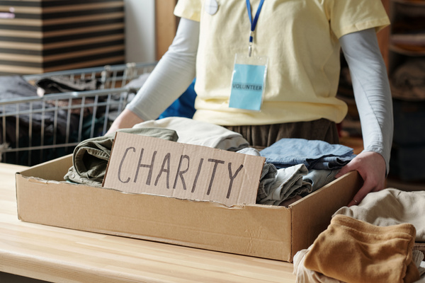 Voluntary worker with blue badge and dressed in pale yellow t-shirt over white longsleeve holds both hands on the charity box with clothes and with cardboard with an inscription about charity which is on the table near with a stack of folded clothes