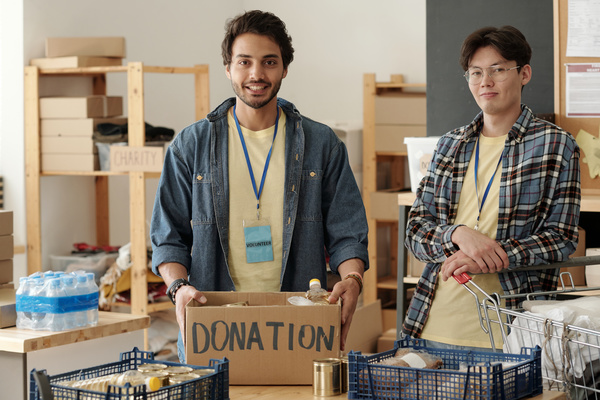 Young smilling male volunteers with dark hairs stand keeping hands on a box filled with products which is on the table near with blue plastic crates with food and cart with donations beside the table