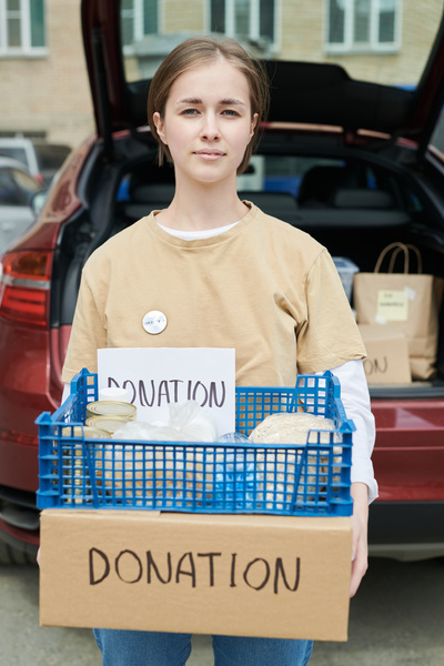 Young female volunteer with short hair wearing white pin badge on her pale yellow t-shirt holds a box and a blue plastic crate on it with donations and a cardboard with a note donation