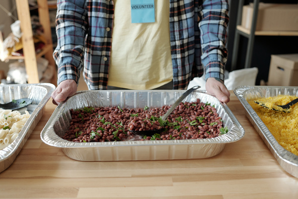 A volunteer in a plaid shirt over a light-coloured T-shirt with two hands holds an aluminum form with beans which is on a table with aluminum form full of polenta and pasta