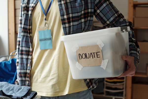 Voluntary worker dressed in checkered shirt over a pale yellow t-shirt holds plastic box with donations with an inscription donate on it