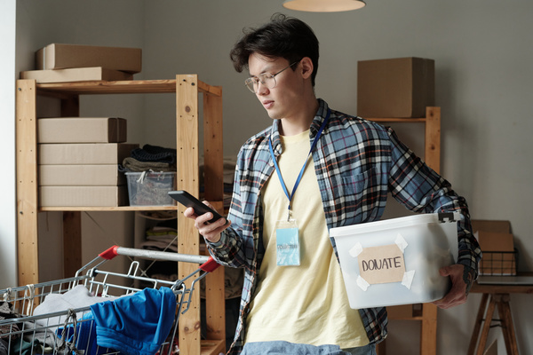 Male dark haired volunteer with glasses and a blue badge in checkered shirt over a pale yellow t-shirt with smartphone in his hand holds plastic box with a note donate on it