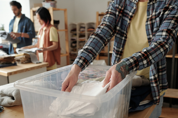 Volunteer in plaid shirt over t-shirt with colored tattoo on his left hand puts folded white item of clothing in plastic box on the table next to which are stacks of folded clothes