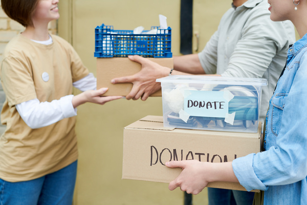A volunteer takes a plastic crate and box from a man standing next to a woman who is holding a box and a plastic container labeled donate and donation that contains items for charity.