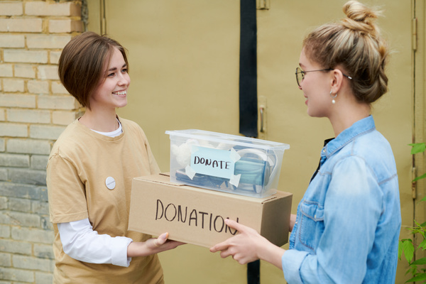 A woman with glasses and in denim shirt with both hands passes a box and a plastic container with donations to the brown - haired volunteer with bob haircut dressed in yellow t-shirt with a white pin badge