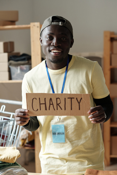 Smiling volunteer with a dark cap dressed in light yellow t-shirt stands with cardboard sign with an inscription charity