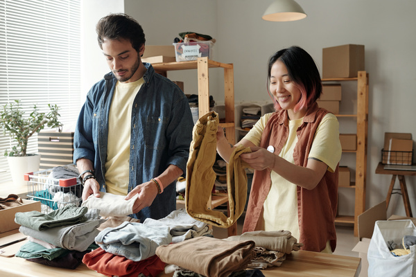 Young smiling voluntary workers in pale yellow t-shirts fold pieces of clothing and stack then on the table with piles of folded pieces of clothing