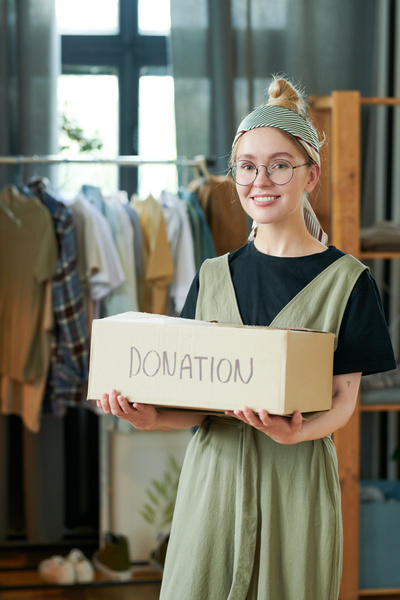 Smiling blonde woman with a bun hairstyle and a headband dressed in swamp colored sleeveless dress over black t-shirt holds box with donations