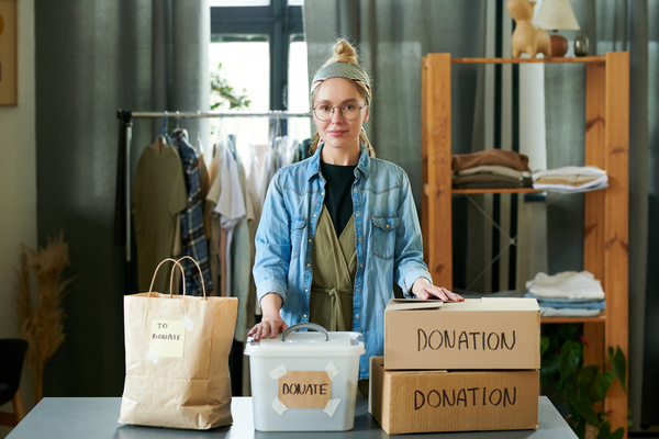 A woman with a bun hairstyle a headband and round glasses dressed in denim shirt keeps her hands on charity box and plastic container with inscriptions donate and donations which are on the table with a craft bag contained stuff to donate