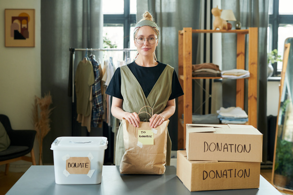 A blonde woman with tied up hair and round glasses dressed in attire of dark colors keeps her hands on craft package with donations which is on the table near between boxes and a plastic container with inscriptions donation and donate
