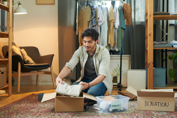 A young man with dark hair and beard dressed in shirt and jeans packs an artickle of clothing in a box with donations sitting on the carpet in the room with clothes rail and dark colored armchair with wooden legs and pillows