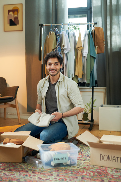 A smiling black-haired man with a beard dressed in a beige shirt jeans packs an item of clothing into a box with donations sitting on ta carpet in a room with a boxes and a clothes rail and paintings on the wall and on the floor