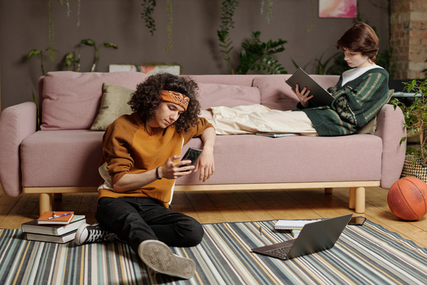 A guy with curly hair in a youth outfit is sitting on the floor surrounded by books and gadgets with a phone in his hands leaning on a pink sofa on which a girl with a short haircut is sitting and reading a book