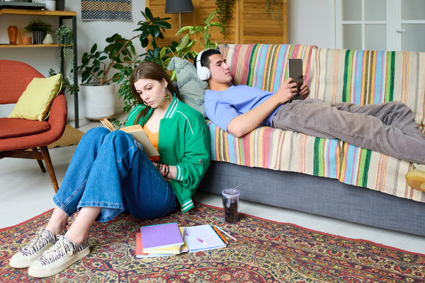 A young girl in a bright outfit is reading a book sitting on the floor next to notebooks near the sofa on which a guy in white headphones is lying with a tablet in his hands