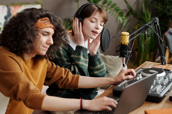 A zoomer girl with short dark hair in a green sweater sings sitting next to a man in a mustard-colored sweatshirt and a banana who uses a laptop and a DJ console