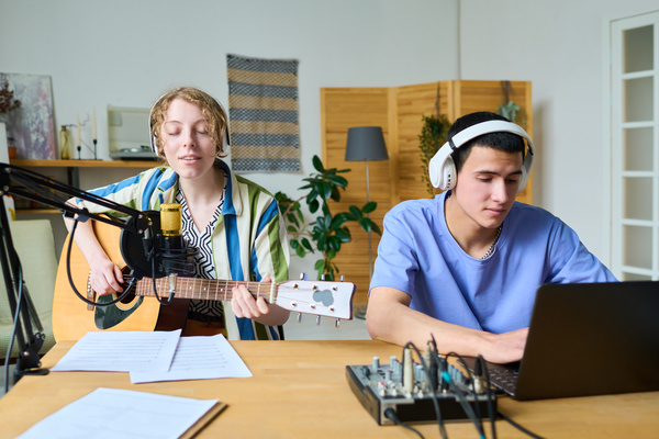 A girl with blonde wavy hair sings and plays guitar into a microphone sitting at a table next to a young man in headphones who is recording music