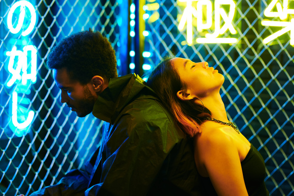 A woman with a short haircut and fashionable accessories sits with her back to a man in a black jacket her head tilted back on his back against an urban background with a metal mesh fence and neon yellow and blue hieroglyphics
