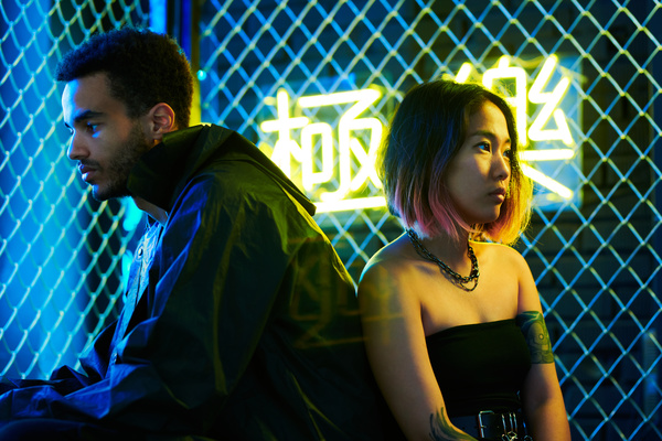 A young man with dark curly hair wearing a black jacket and a woman with a short haircut and tattoos and stylish accessories sit with their backs to each other on an urban background with a metal mesh fence and neon yellow and blue hieroglyphs
