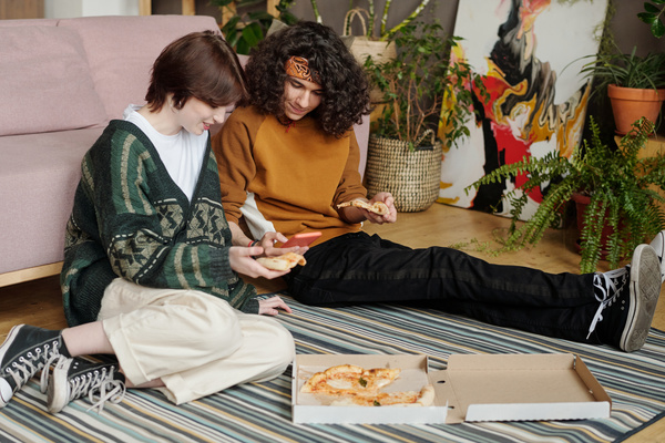 A girl with a pixie haircut holding a piece of pizza in her hands sits with her legs bent on a striped carpet next to a curly-haired guy in a bandana who takes a photo of a pizza box lying nearby on the floor
