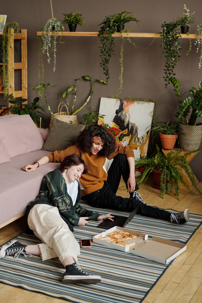 A young woman with short hair reclining on a striped carpet laughs while looking at a laptop with a smiling guy with curly hair sitting next to her next to whom there is a pizza box in a room with plants