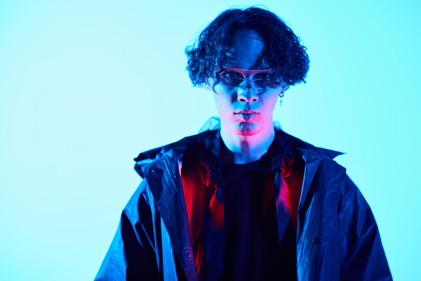 A close-up of a cyberpunk man with wavy hair wearing earring in the form of a cross and dark clothes who is lightened with blue and red neon light