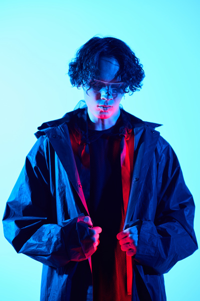 A man with wavy dark hair in a black raincoat with red neon light on inside holds the edges of it with both hands stands against a light-colored background looking down
