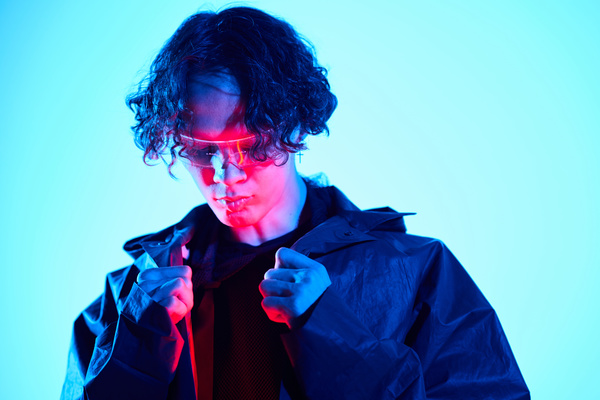 The face of a young man with curly hair illuminated with red neon light who holds the collar of his raincoat with his hands