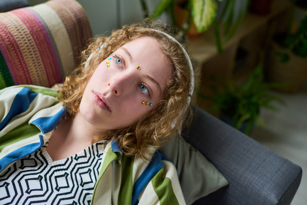 A zoomer girl with light curly hair and blue eyes wearing headphones is lying on the couch dreamily looking away