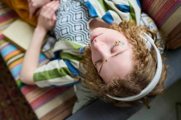 A curly blonde with yellow and blue rhinestones on her face in a colorful outfit with prints in white headphones lies on the couch with her eyes closed