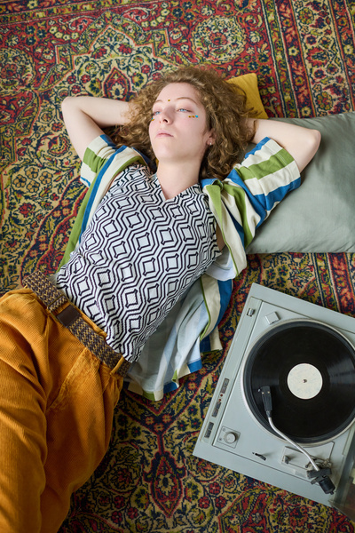 A young girl with curls in bright clothes is lying on the carpet with her hands behind her head next to the gramophone thoughtfully looking away