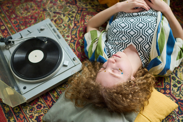 A girl with curly hair lies on the carpet with her hands folded on her stomach and listens to music from a gramophone with her eyes closed