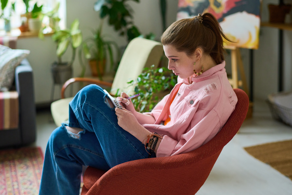 A young girl with a ponytail in a pink denim jacket and jeans with holes is sitting in a red armchair and making notes in a notebook